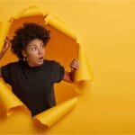 National Arts Fundraising School - Woman poking head through paper tear hole on orange background.